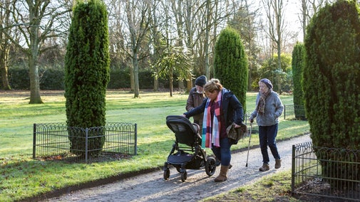 A multi-generational family of three adults and one child walk through the gardens at Speke Hall. They are dressed for winter and pushing a pram.